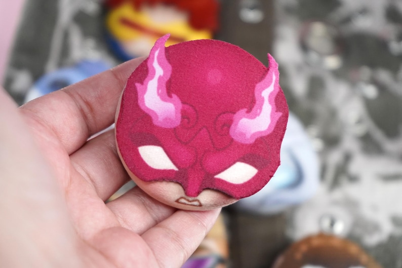 Hand holding a plush head pin badge button featuring a male character in a red face mask with horns.