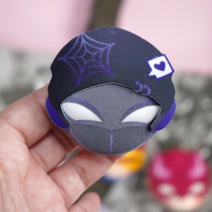 Hand holding a plush head pin badge button featuring a male character dressed in a black outfit and a hat.