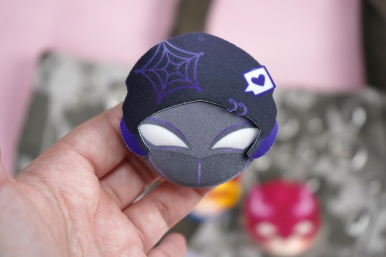 Hand holding a plush head pin badge button featuring a male character dressed in a black outfit and a hat.