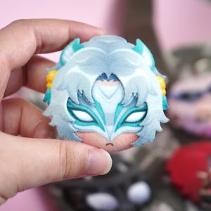 Hand holding a plush head pin badge button featuring a female fox character with white hair and a face mask as well as green ears.