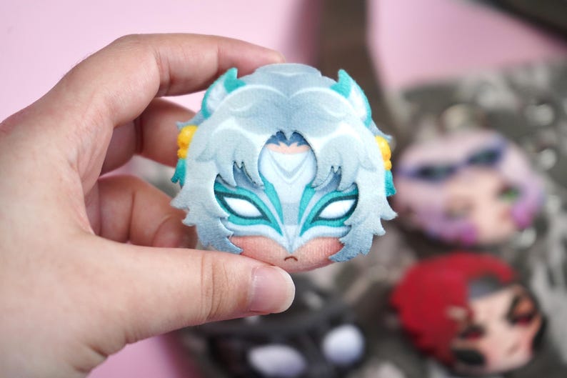 Hand holding a plush head pin badge button featuring a female fox character with white hair and a face mask as well as green ears.