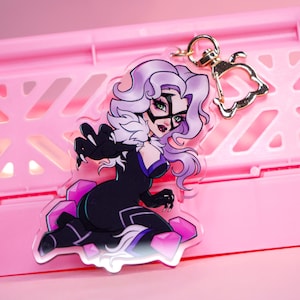Puede incluir: Un llavero acrílico transparente con una ilustración estilizada de un personaje con un catsuit negro con ribete de piel blanca y una máscara. El personaje tiene el pelo morado y está posando sobre una base geométrica rosa y morada. El llavero tiene un cierre dorado.