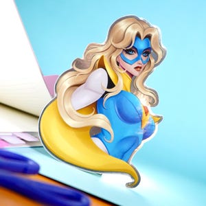 May include: A die-cut sticker featuring a female superhero with long blonde hair, a blue mask, and a blue and yellow costume. The sticker is set against a light blue background, with a notebook and pens visible.
