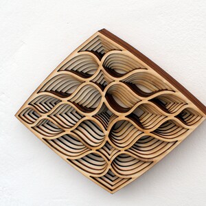 May include: A wooden wall art piece with a geometric design of overlapping, layered curves. The piece is made of light brown wood and has a diamond shape.