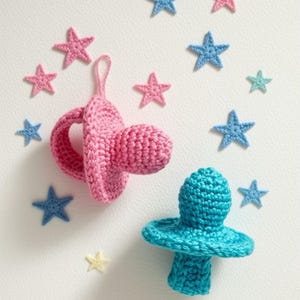 Op de afbeelding: Twee gehaakte fopspenen, één roze en één turquoise, met sterdecoraties. De afbeelding toont roze, blauwe en een enkele gele gehaakte ster op een witte achtergrond. Baby shower thema.