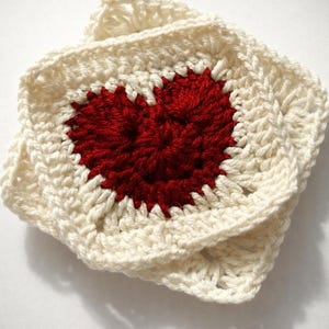Può includere: Quadrato all'uncinetto con un cuore rosso al centro. Il cuore è circondato da un bordo color crema. L'articolo fatto a mano è probabilmente un sottobicchiere o un elemento decorativo.