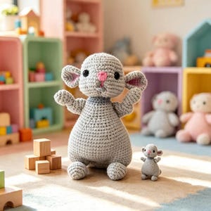 Può includere: Un grande giocattolo a forma di topo lavorato all'uncinetto in grigio con naso rosa e occhi neri, in piedi accanto a un topo all'uncinetto molto più piccolo. Entrambe le figure hanno orecchie arrotondate, braccia corte e una superficie in filato.