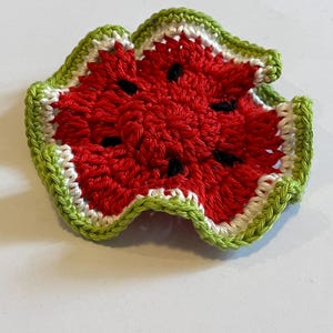 Può includere: Un sottobicchiere rotondo all'uncinetto a forma di fetta di anguria, con un centro rosso vivace, semi neri ricamati, un sottile anello bianco e un bordo smerlato verde chiaro. Il pezzo in filato presenta un bordo ondulato e arricciato.