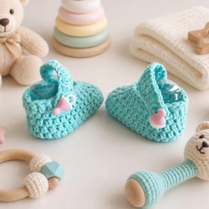 Può includere: Un paio di scarpine per neonati all'uncinetto color azzurro, con un piccolo bottone a forma di ciuccio rosa sul lato di ogni scarpa. Le scarpine sono circondate da vari giocattoli per neonati, tra cui un orsacchiotto e un sonaglio in legno.