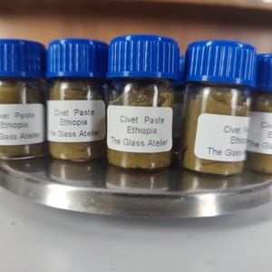Peut inclure: Plusieurs petits pots en verre avec des couvercles bleus, remplis d'une pâte vert-brun. Chaque pot porte l'étiquette "Civet Paste Ethiopia The Glass Atelier". Les pots sont disposés sur un plateau argenté.
