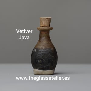Puede incluir: Pequeña botella de vidrio hecha a mano con tapón de corcho. La botella es de color marrón oscuro en la base, pasando a un marrón más claro y rematada con un corcho beige. El texto "Vetiver Java" es visible.