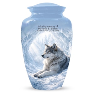 May include: A light blue cremation urn with a wolf illustration. The urn has a lid and features the text "In loving memory of Marlowe E. Sutton" and dates. The wolf is depicted in a snowy landscape.
