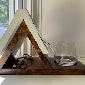 May include: A wooden triangular stand with a book holder on top and a glass holder on the bottom. The stand is dark brown and has a natural wood finish. A pair of glasses is resting on the stand.