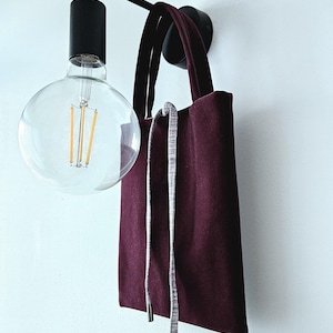 Puede incluir: Una bolsa de mano color burdeos con un cordón de ajuste gris que cuelga de la parte superior. La bolsa está colgada de una lámpara de pared negra con una bombilla.