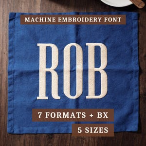 May include: A square blue fabric napkin featuring the cream-colored embroidered name ROB in a bold serif font. Text overlays include MACHINE EMBROIDERY FONT, 7 FORMATS + BX, and 5 SIZES in brown rectangular boxes on the blue fabric surface.