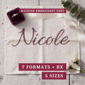 Peut inclure: Un carré de tissu crème présentant le prénom Nicole brodé à la machine en écriture mauve foncé. Une boîte à bague violet foncé avec une bague argentée est placée dans le coin, accompagnée d'un ruban blanc et de deux boutons de rose blancs.