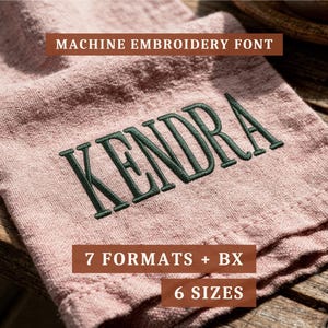 May include: A dusty pink linen fabric featuring the dark green embroidered name KENDRA in a serif font. Text overlays state MACHINE EMBROIDERY FONT, 7 FORMATS + BX, and 6 SIZES. The fabric has a visible woven texture and is folded on a wooden surface.