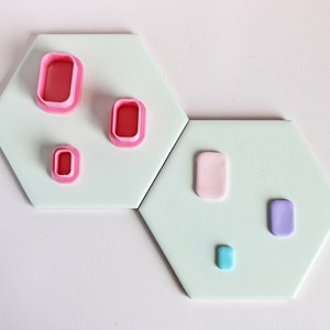 May include: Five different sized and coloured rectangular shapes made of polymer clay on a light green background. The shapes are pink, light pink, light blue, and purple.