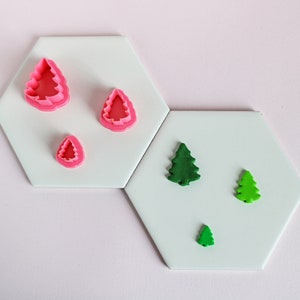 Peut inclure: Trois emportes-pièces en plastique en forme de sapin de Noël, deux roses et trois verts, sur une surface blanche. Les emportes-pièces sont disposés par ordre de taille, le plus grand en haut et le plus petit en bas.