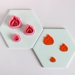 May include: A selection of pink flame-shaped cookie cutters and orange flame-shaped fondant decorations are displayed on hexagonal tiles. The cutters are nested, showing different sizes. The fondant flames are in various sizes.