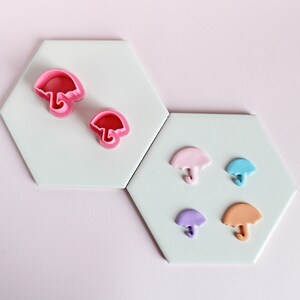 May include: Two pink plastic cookie cutters shaped like umbrellas, and five fondant umbrellas in pink, blue, purple, and orange on a white hexagonal tile surface.