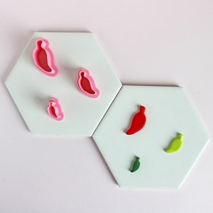 May include: Five different cookie cutters in the shape of chili peppers. The cutters are on a white surface. The cutters are red, green, and pink.