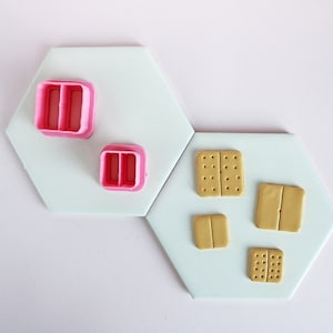 May include: Two pink plastic cookie cutters shaped like a rectangle with two holes in the middle, and four brown clay squares with a line down the middle, on a white hexagonal surface.