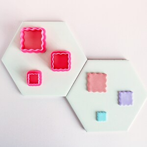 May include: A selection of pink and pastel square cookie cutters with scalloped edges. The cutters are displayed on hexagonal tiles, against a light pink backdrop. The cutters vary in size, with the largest being approximately 5 cm square.