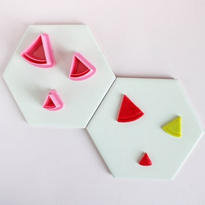 Peut inclure: Cinq emportes-pièces en plastique en forme de tranches de pastèque. Les emportes-pièces sont roses, rouges et jaunes. Les emportes-pièces sont disposés sur deux carreaux hexagonaux blancs.