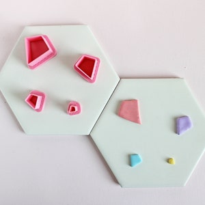 May include: A set of pink and purple fondant cutters in various shapes, including diamonds, hearts, and a star. The cutters are arranged on two white hexagonal tiles.