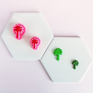 May include: Two pairs of palm tree-shaped polymer clay cutters. One pair is bright pink, and the other is green. The cutters are placed on white, hexagonal tiles against a light pink background. These are likely for crafting or baking.