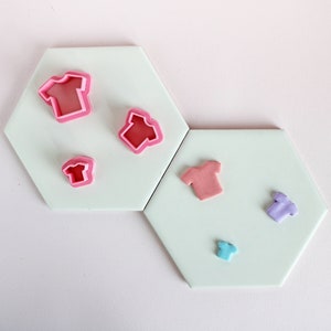 May include: Five different sized pink, red and purple plastic cookie cutters shaped like t-shirts on a white hexagonal surface.