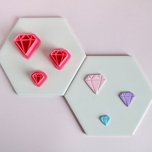 Puede incluir: Cinco cortadores de galletas en forma de diamante sobre un fondo blanco. Los cortadores son de color rosa, morado claro y azul. Los cortadores están dispuestos en un patrón de tamaño decreciente.