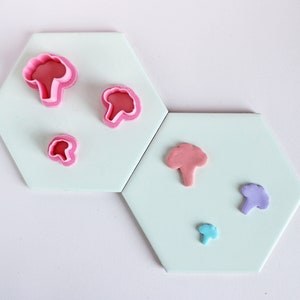 May include: Five pink and purple fondant cutters shaped like broccoli florets on a white background. The cutters are arranged in a random pattern on two white hexagonal tiles.