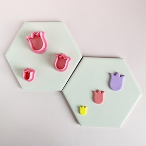 Peut inclure: Cinq emportes-pièces en forme de tulipe de différentes tailles, de couleur rose et violette, sur une surface blanche. Les emportes-pièces sont disposés en rangée du plus grand au plus petit.