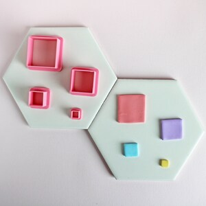 May include: A set of six square cookie cutters in various sizes. The cutters are pink, light blue, purple, and yellow. The cutters are arranged on two white hexagonal tiles.