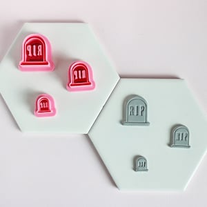 May include: Set of five tombstone-shaped cookie cutters in two sizes. The larger cutters are gray and the smaller cutters are pink. All cutters feature the inscription "RIP".
