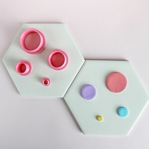 Op de afbeelding: Vijf ronde koekjesvormen in roze en twee in blauw op een lichtgroene achtergrond. De koekjesvormen zijn in een patroon op twee zeshoekige tegels gerangschikt.