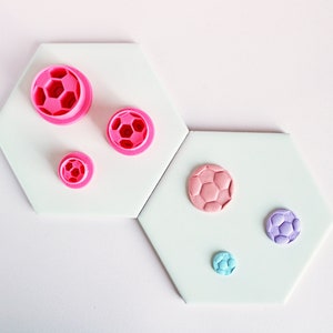 May include: Five different sized fondant cutters shaped like footballs. The cutters are pink, purple, and blue. The cutters are on a white background.