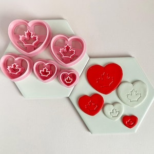 Peut inclure: Ensemble de quatre emporte-pièces en forme de cœur avec un motif de feuille d'érable canadienne. Les emporte-pièces sont de différentes tailles et sont en plastique rose. Il y a aussi trois biscuits en forme de cœur, deux rouges et un blanc, avec le motif de la feuille d'érable.