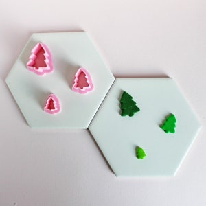 May include: A set of pink and green Christmas tree cookie cutters on a white background. The cutters are arranged in different sizes.