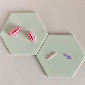 May include: Four plastic cookie cutters in the shape of swords. Two are pink, one is light pink, and one is purple. The cutters are on a light green background.
