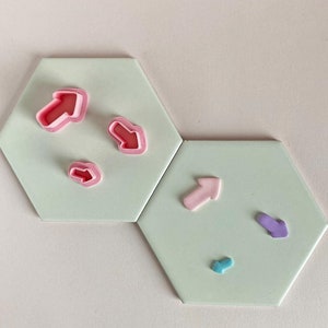 May include: A set of six arrow-shaped cookie cutters in various colors, including pink, purple, and blue. The cutters are arranged on two hexagonal tiles.