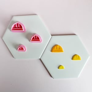 May include: Five different sized plastic cookie cutters in the shape of a hard hat. The cutters are pink and yellow. The cutters are arranged on two white hexagonal tiles.
