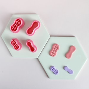 May include: A set of pink peanut-shaped cookie cutters and pink and purple fondant peanut shapes on a light green background.