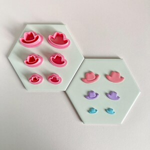 May include: A selection of cowboy hat-shaped cookie cutters in pink, purple, and blue. The cutters are displayed on two hexagonal tiles. The hats are of varying sizes, with larger pink hats and smaller purple and blue hats.