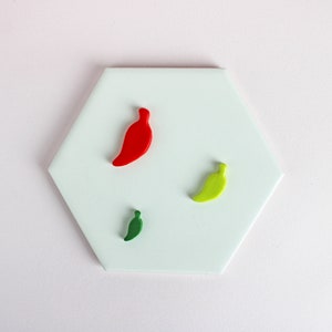 May include: Three ceramic chili pepper ornaments on a white hexagonal tile. One chili pepper is red, one is green, and one is yellow.