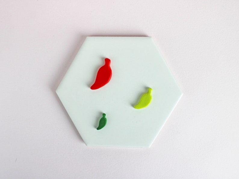 May include: Three ceramic chili pepper ornaments on a white hexagonal tile. One chili pepper is red, one is green, and one is yellow.