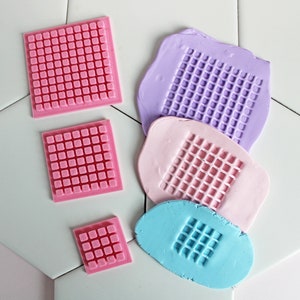 May include: Four square silicone molds with a grid pattern. The molds are pink, purple, light pink, and blue.