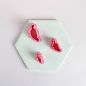 May include: Three pink plastic cookie cutters shaped like chili peppers on a light blue hexagonal surface.